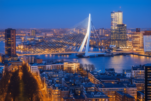 Twilight view of Rotterdam featuring the Erasmus Bridge illuminated over the Nieuwe Maas River, with the modern city skyline and historic buildings in view.